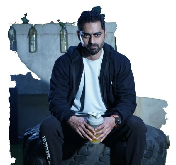 Indian athlete sitting on a tire in a gym setting, wearing a The Toxic Fitness black tracksuit and white sneakers, holding a shaker bottle, with gym equipment in the background.